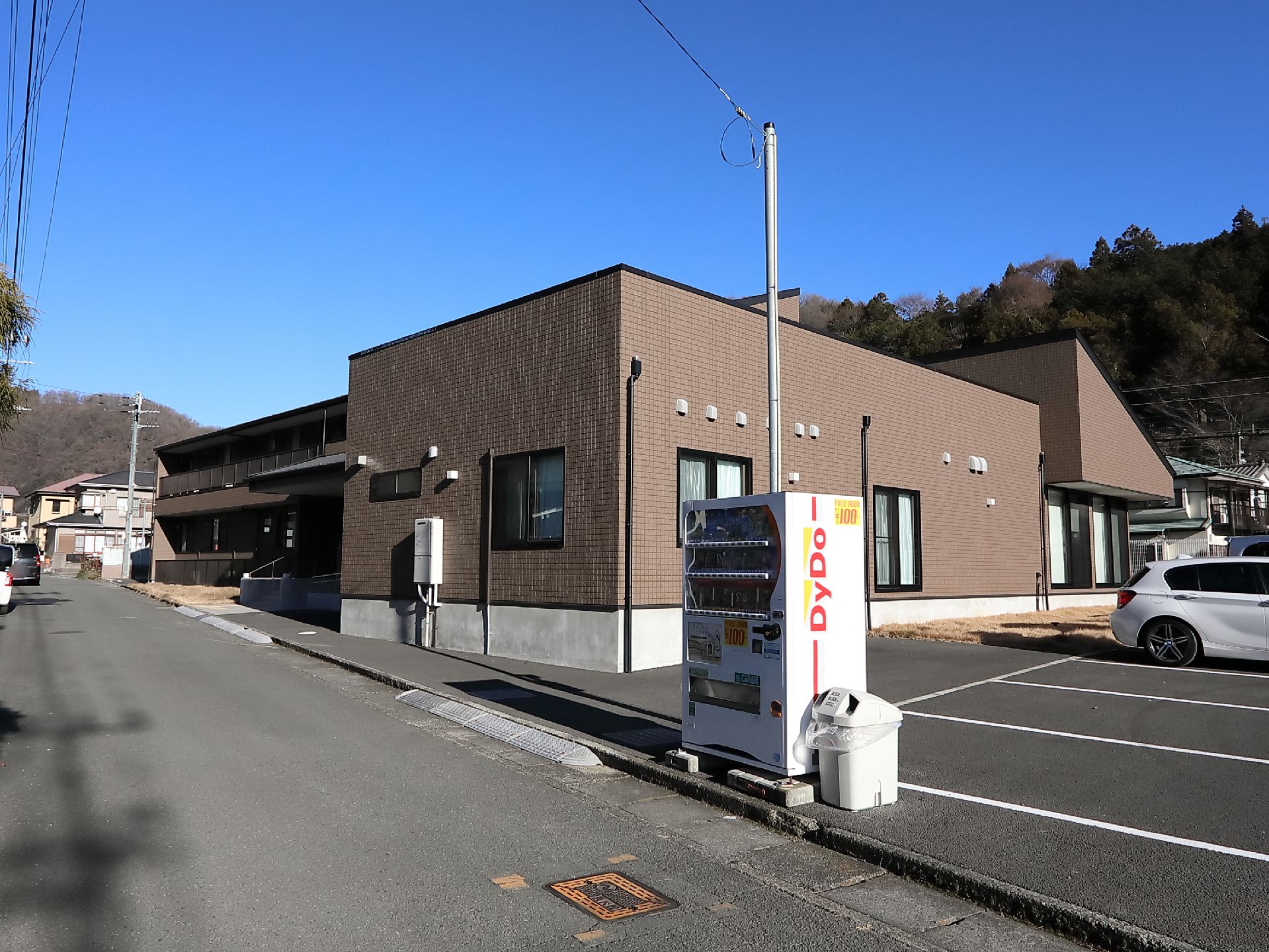 道路に面したところに施設建物があり、隣接して駐車場がある。道路からの乗り入れ部には段差をなくすプレートがある。