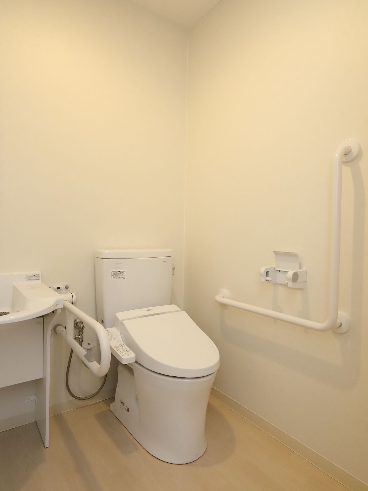 トイレの角に温水洗浄便座があり、隣に洗面台がある。便座の横の壁にL字の手すりとペーパーホルダーがある。