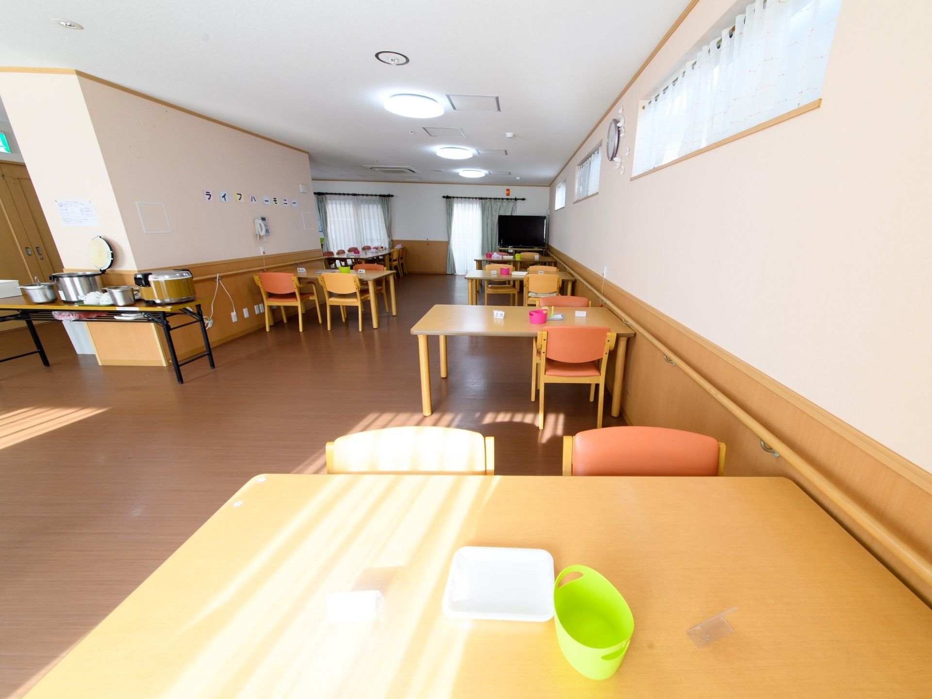 机と椅子が並べられている。窓から明るい日差しが差し込み、掃除も行き届いた綺麗な食堂。壁には手すりが設置されている。