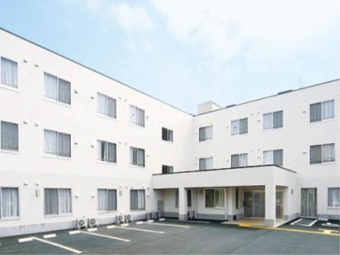 広い駐車場と白い壁の３階建てのL字状になっている建物