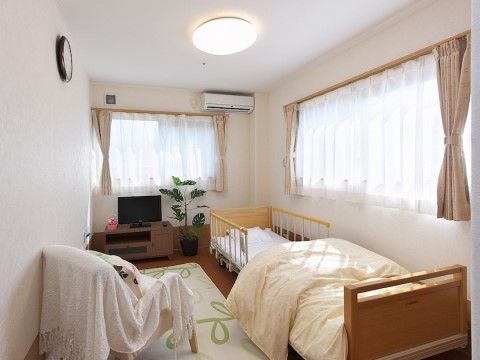 窓から光があふれる清潔感のある居室。ベッドや布カバーがかけられた椅子、テレビや観葉植物が置かれている。