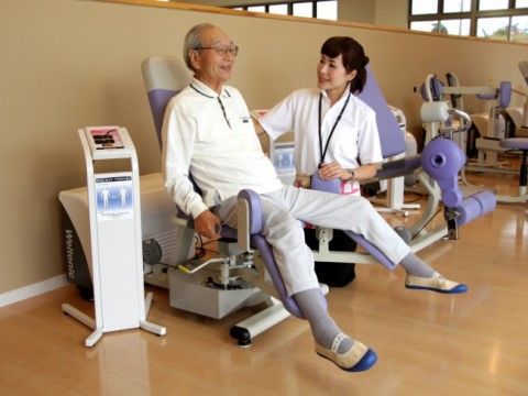 エクササイズマシンに座って運動する入居者と、膝立ちになり目線を合わせてサポートするスタッフ