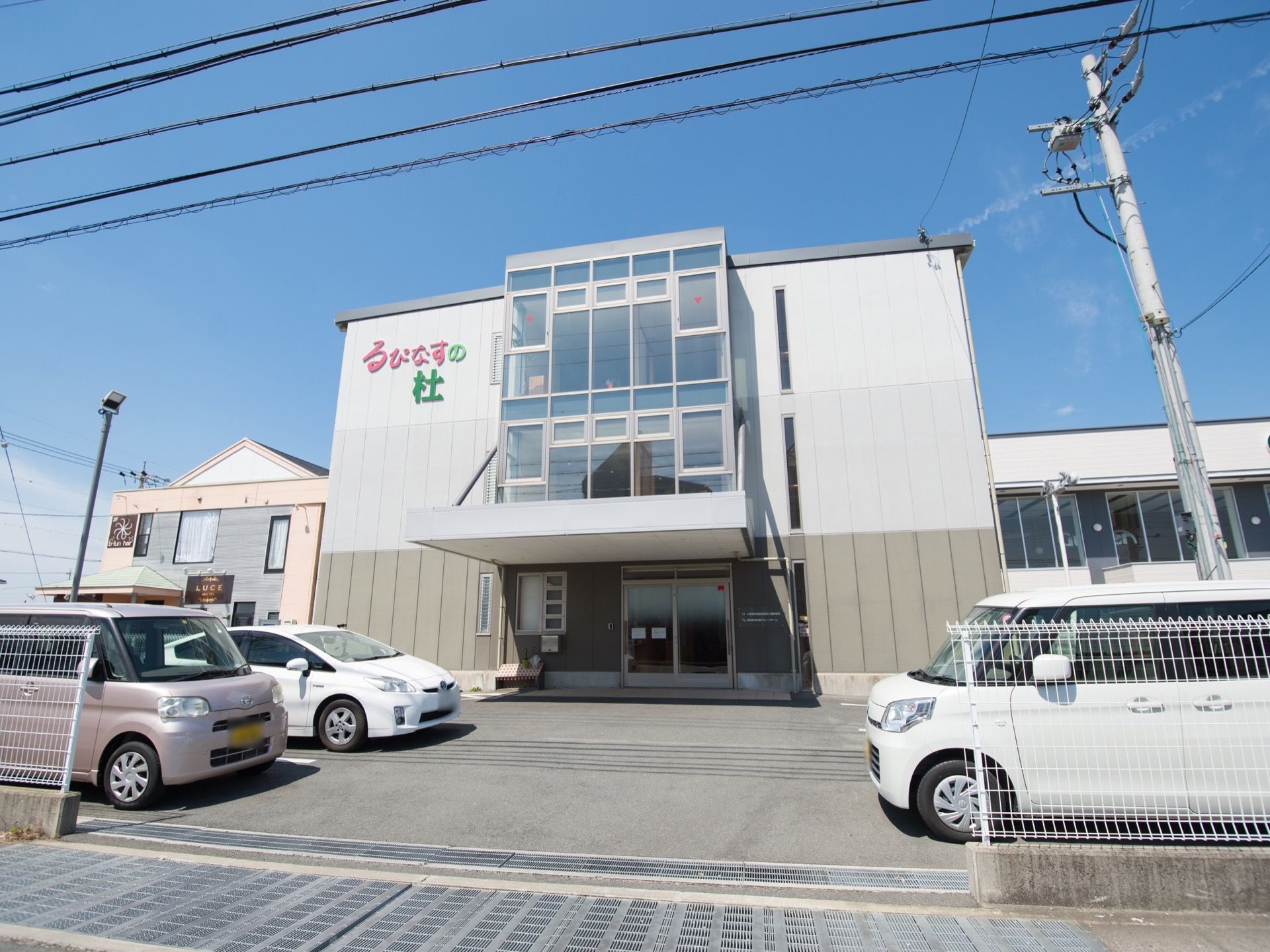 白とグレーに塗られた３階建ての建物で、施設の前には駐車スペースが広がっている。自動車が３台停められている。