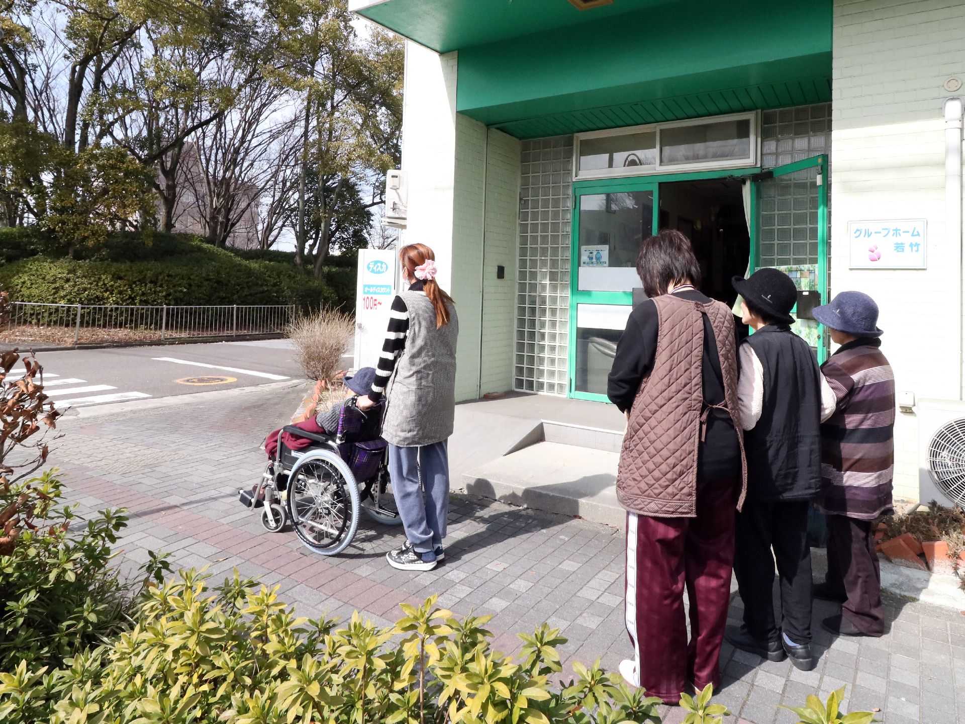 ヘルパーに車椅子を押され、敷地外に出る入居者と、見送るヘルパーと入居者の方々