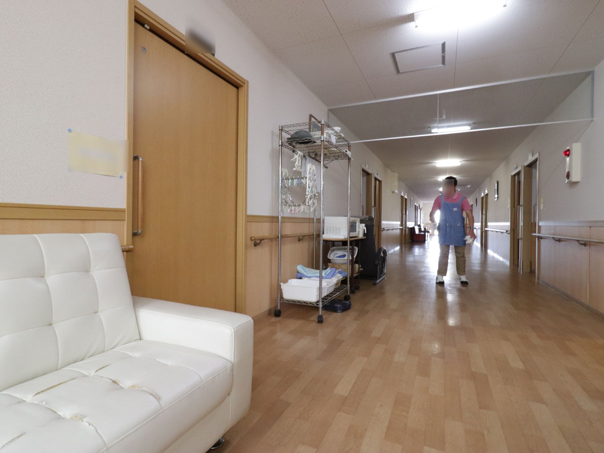 白い大きなソファーが、通路の左端に置いてある。ベージュ色のチノパンにピンク色のシャツという服装のスタッフが、真ん中に立っている。