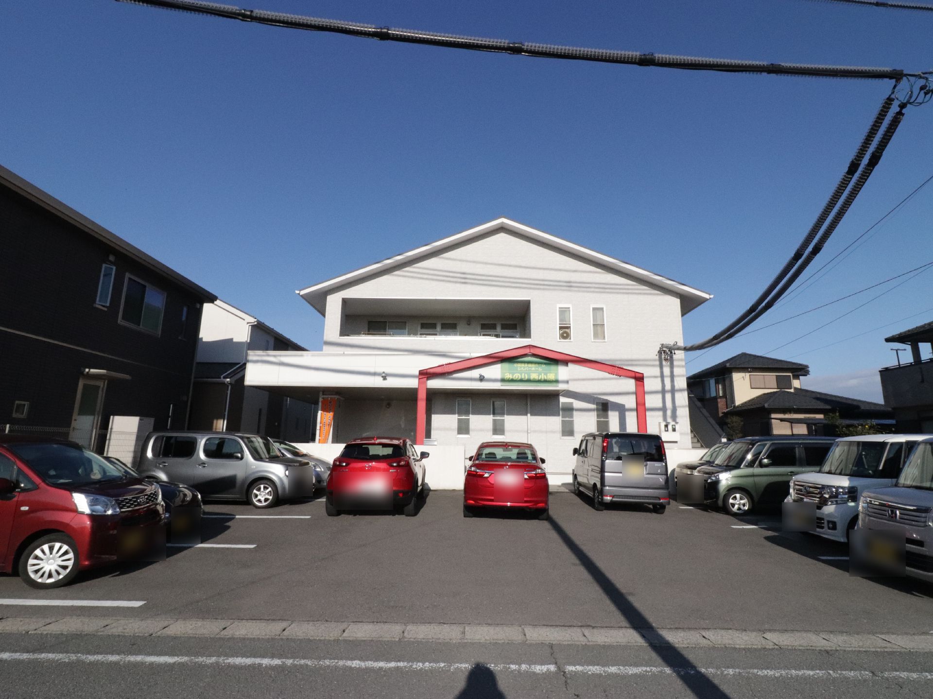 広々とした駐車スペースには、赤い普通自動車など数台の車がコの字型に並んで置いてある。上空には、太い電線が見える。