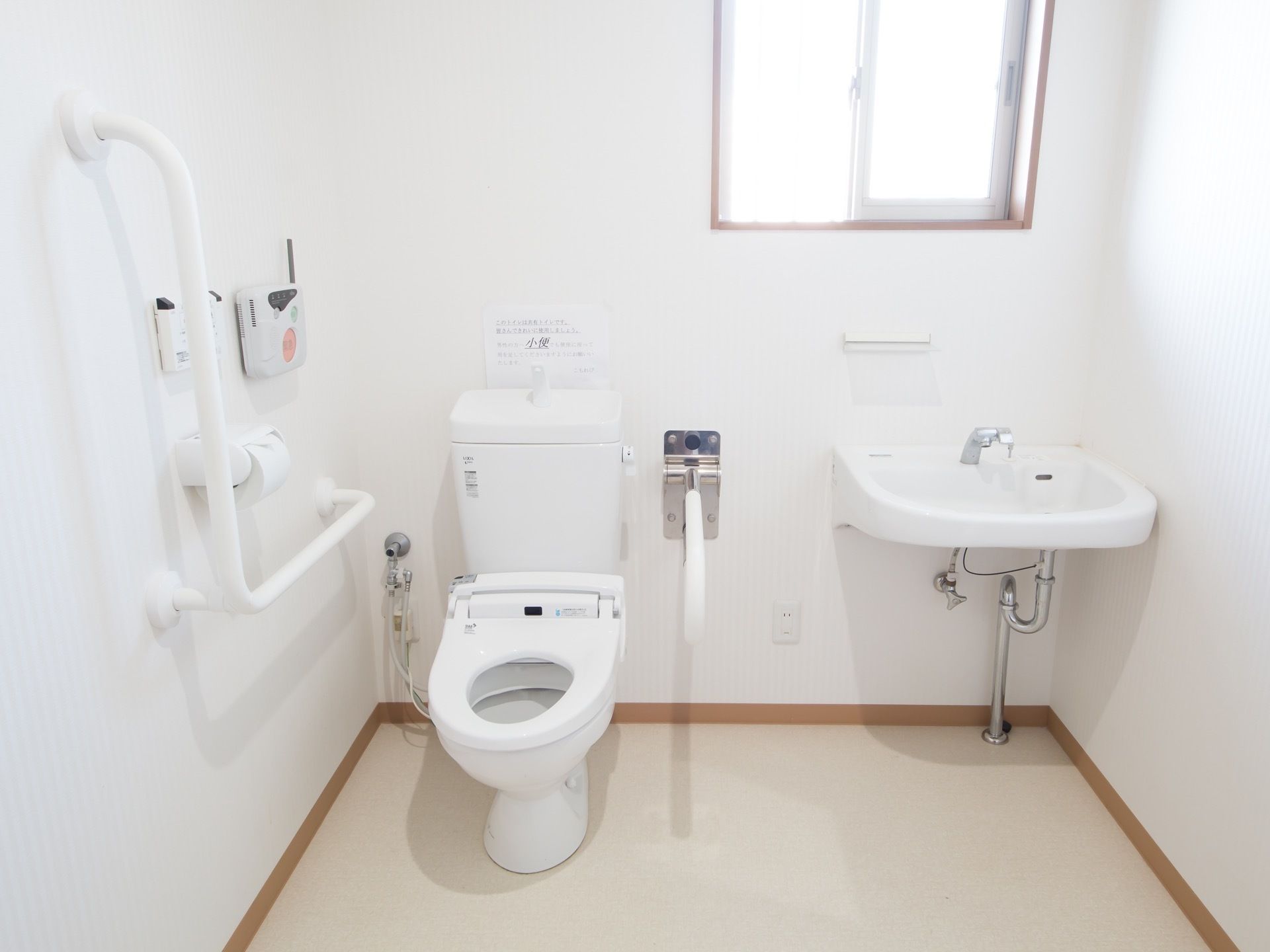 トイレには温水洗浄便座と、ボウルが大きめの手洗いが並んでいる。便器の横と壁には手すりがある。壁に緊急ボタンがある。
