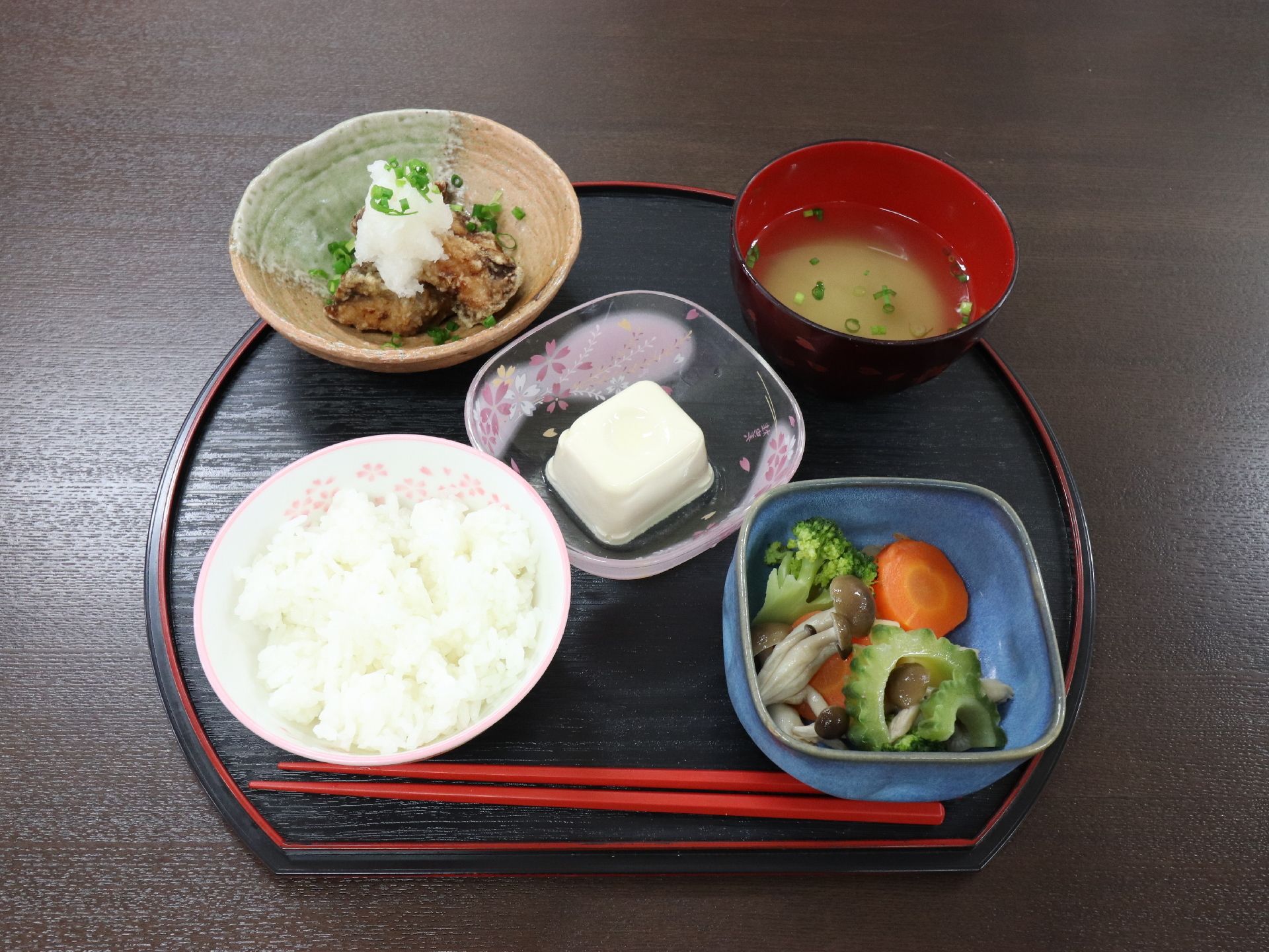 黒いトレーの上に1人分の食事がセットされている。一番手前に箸が置かれており、ご飯と味噌汁のほか、おかず3品が並んでいる。