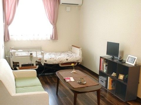 出窓があり、介護用のベッド、エアコン、テレビ、机にゆったりとくつろげるソファーが設備されている居住空間