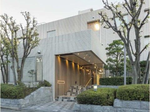 石造りのエントランスに木が数本植えられ、建物の壁面が白いタイル張りの外観