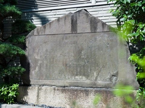 灰色の建物の前に、大きな石でできた碑が設置されており、左右には木が生い茂っている。石の様子から歴史を感じることができる。