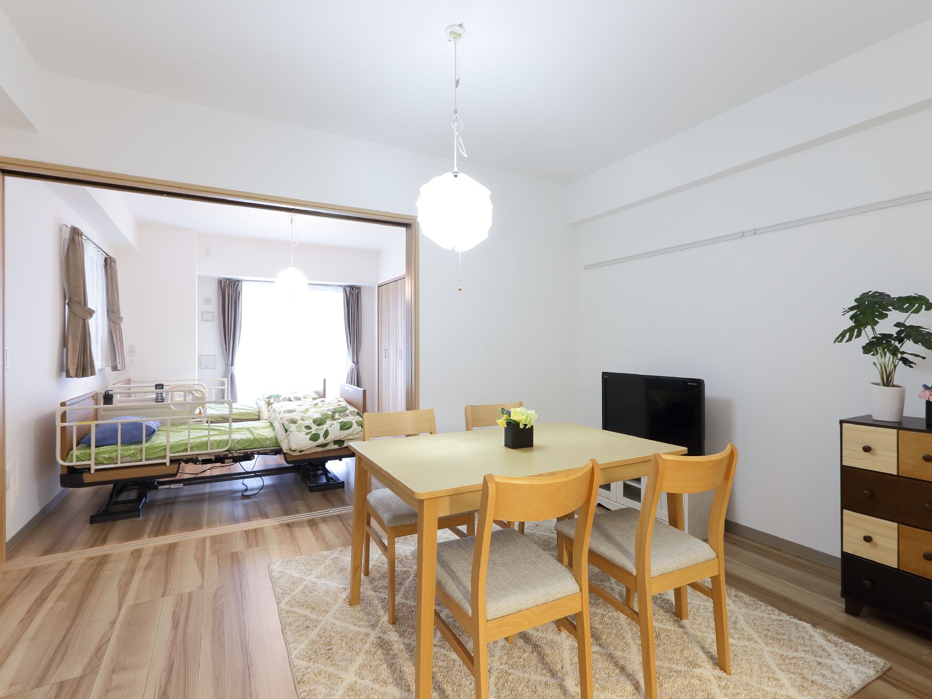 球体の照明器具が天井からぶら下がっている。手前には4つの椅子にテーブルがある。テーブルの右手にテレビが置いてある。奥に寝室がある。