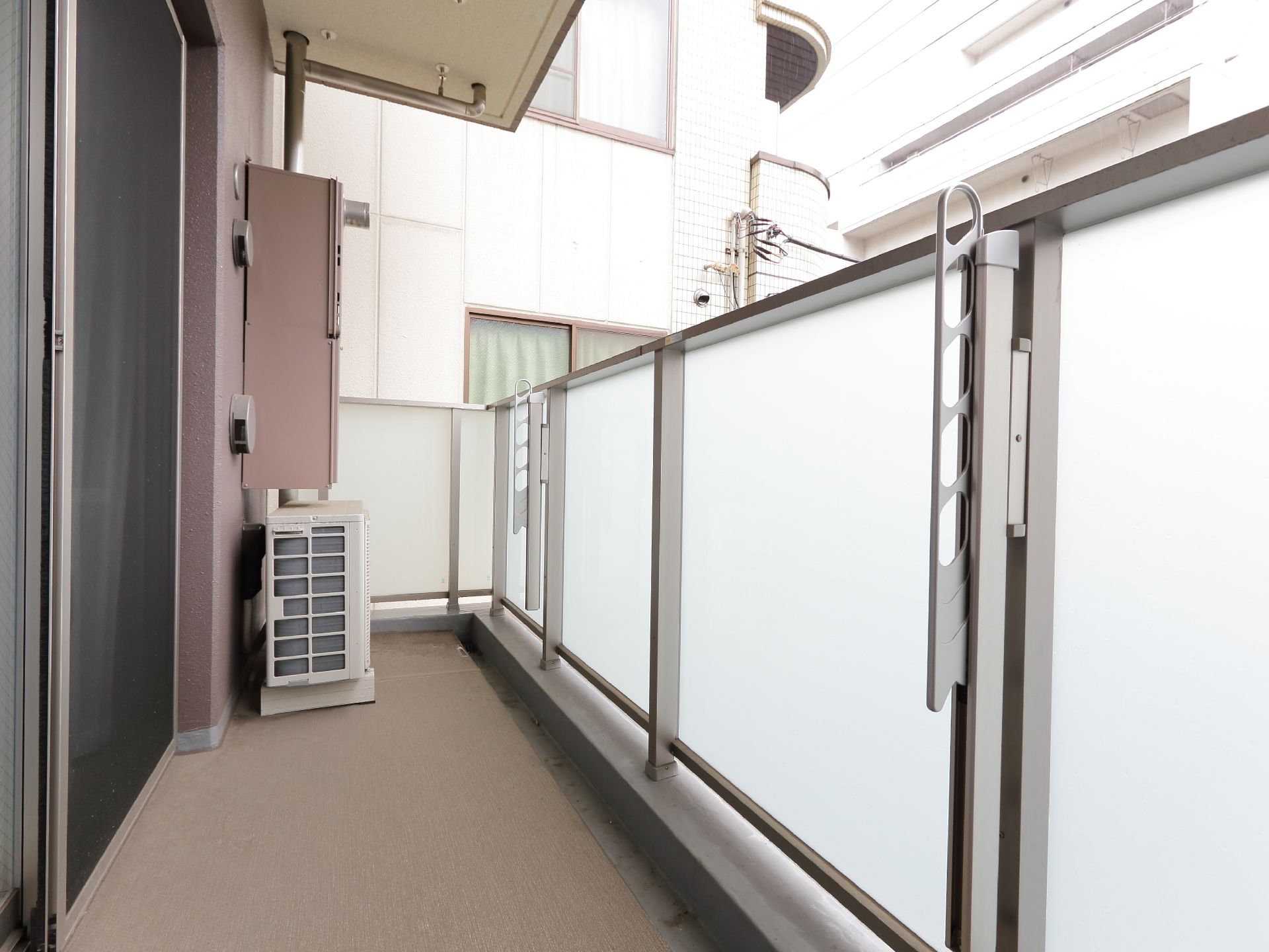 屋根が少しせり出している。左側には窓がある。室外機が室内側に寄せて置いてある。柵は白濁色になっている。