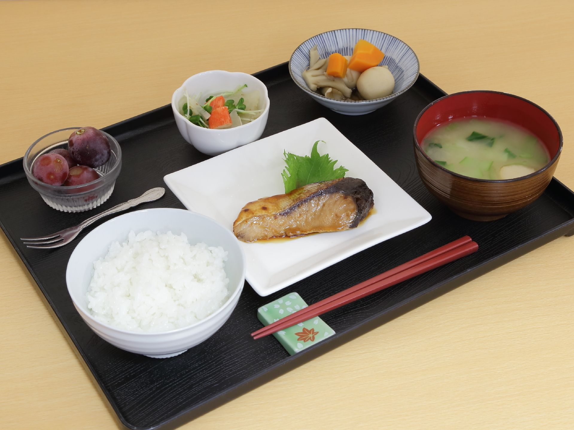 お盆の上に箸が一膳ある。ご飯が茶碗1杯ある。主食に魚があり、味噌汁が付いている。2品の副菜と果物が付いている。