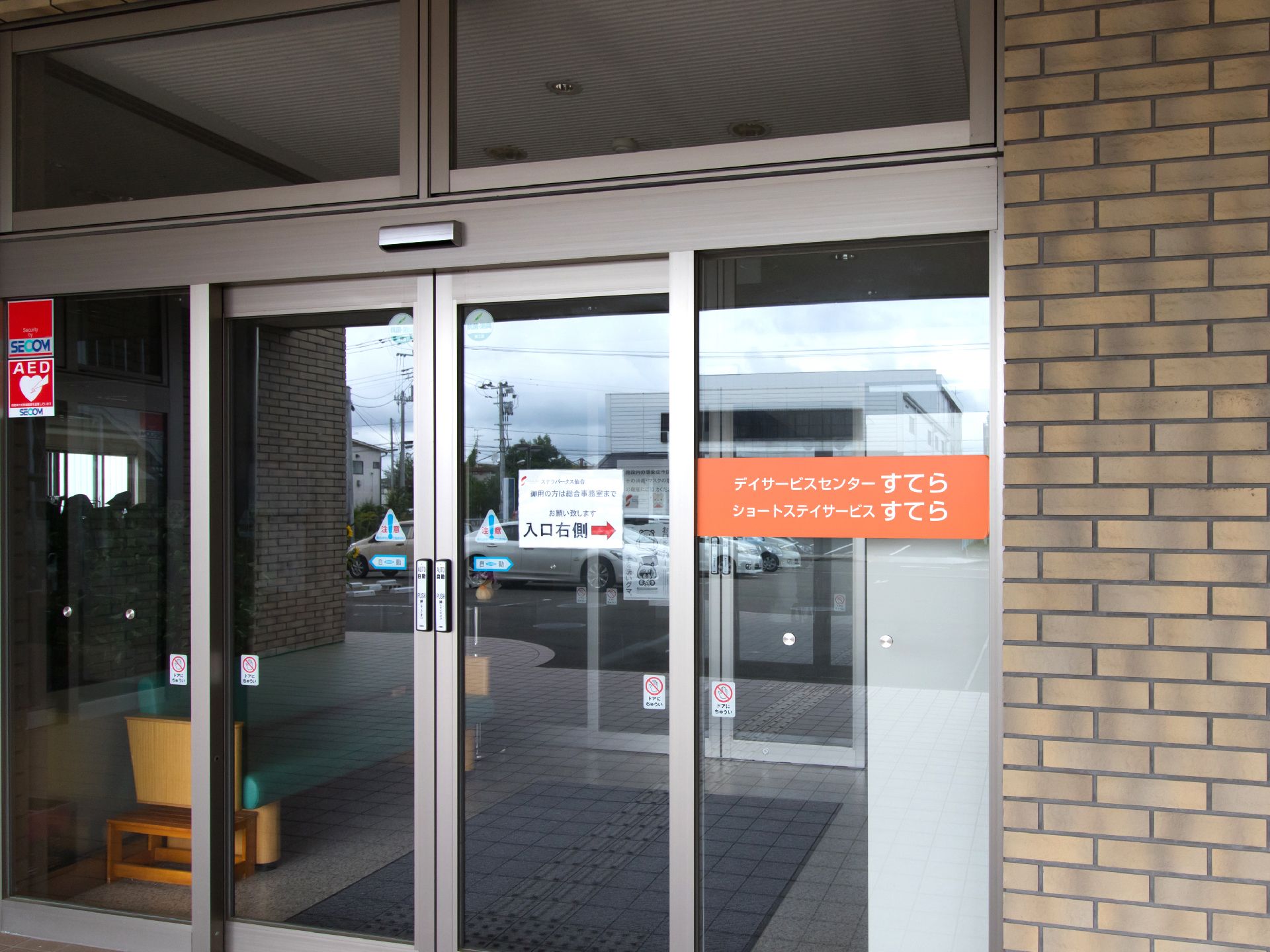 施設の入り口はガラス張りの自動ドアで内部の様子が見える。右側のガラス面にはオレンジ地に白い文字で施設名が書かれている。