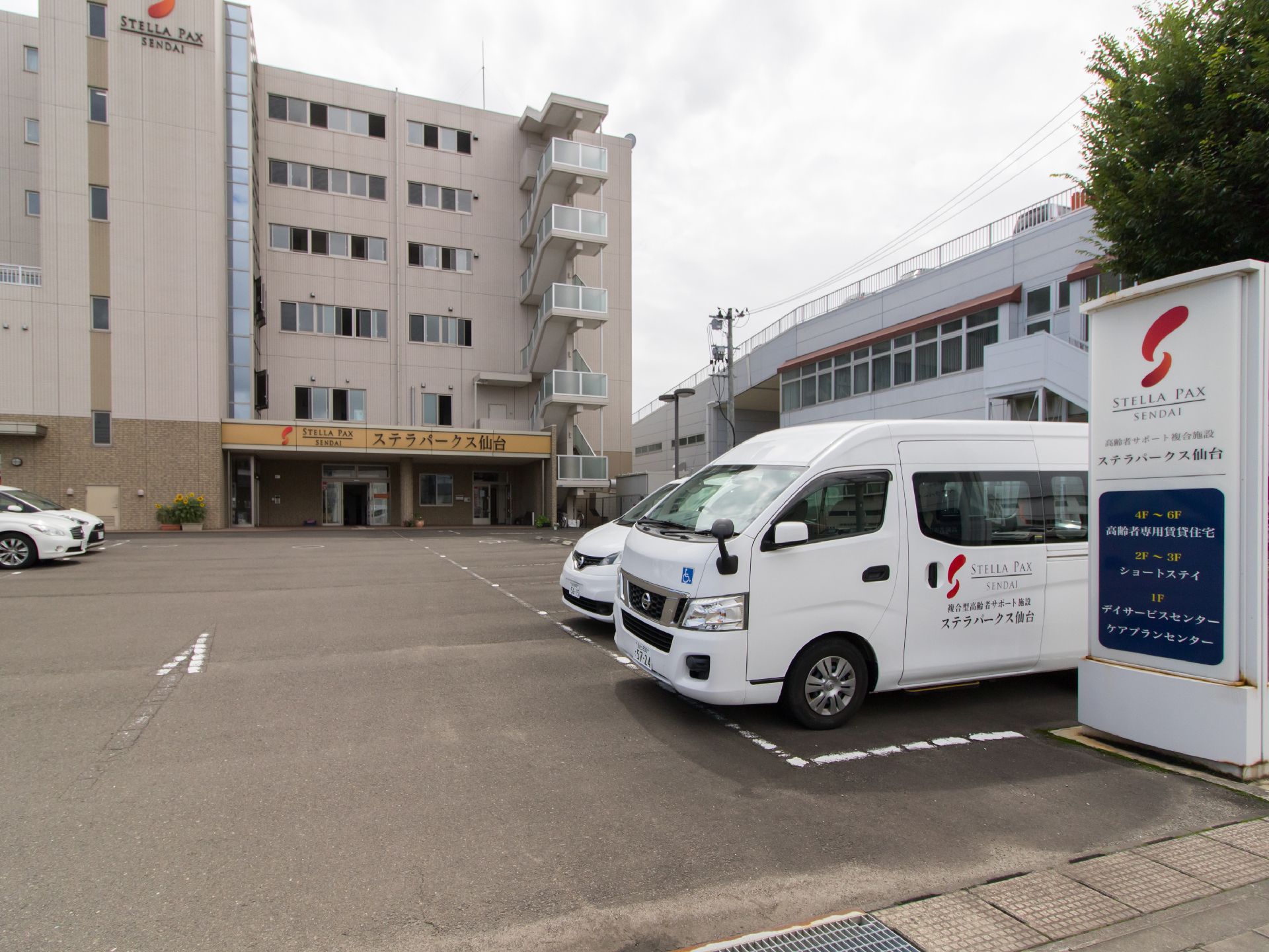 施設は6階建てのマンションで、外壁は明るいベージュである。前面には広い駐車場があり、入り口付近に施設名の入った車が止まっている。