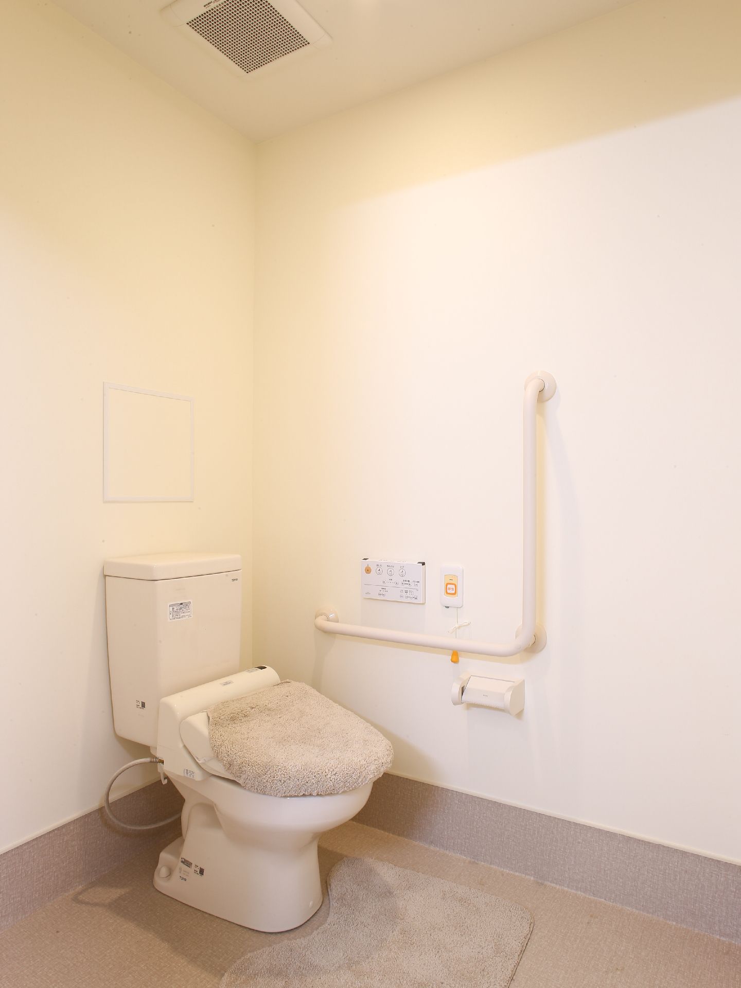 トイレの座面にはカバーがついている。床にはマットが敷いてある。壁面には手すりがついている。呼び出しボタンもある。