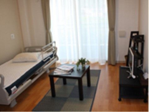 掃き出し窓のあるフローリングの室内に、電動介護ベッドやテーブル、テレビなどが置かれている。壁にはナースコールが設置されている。