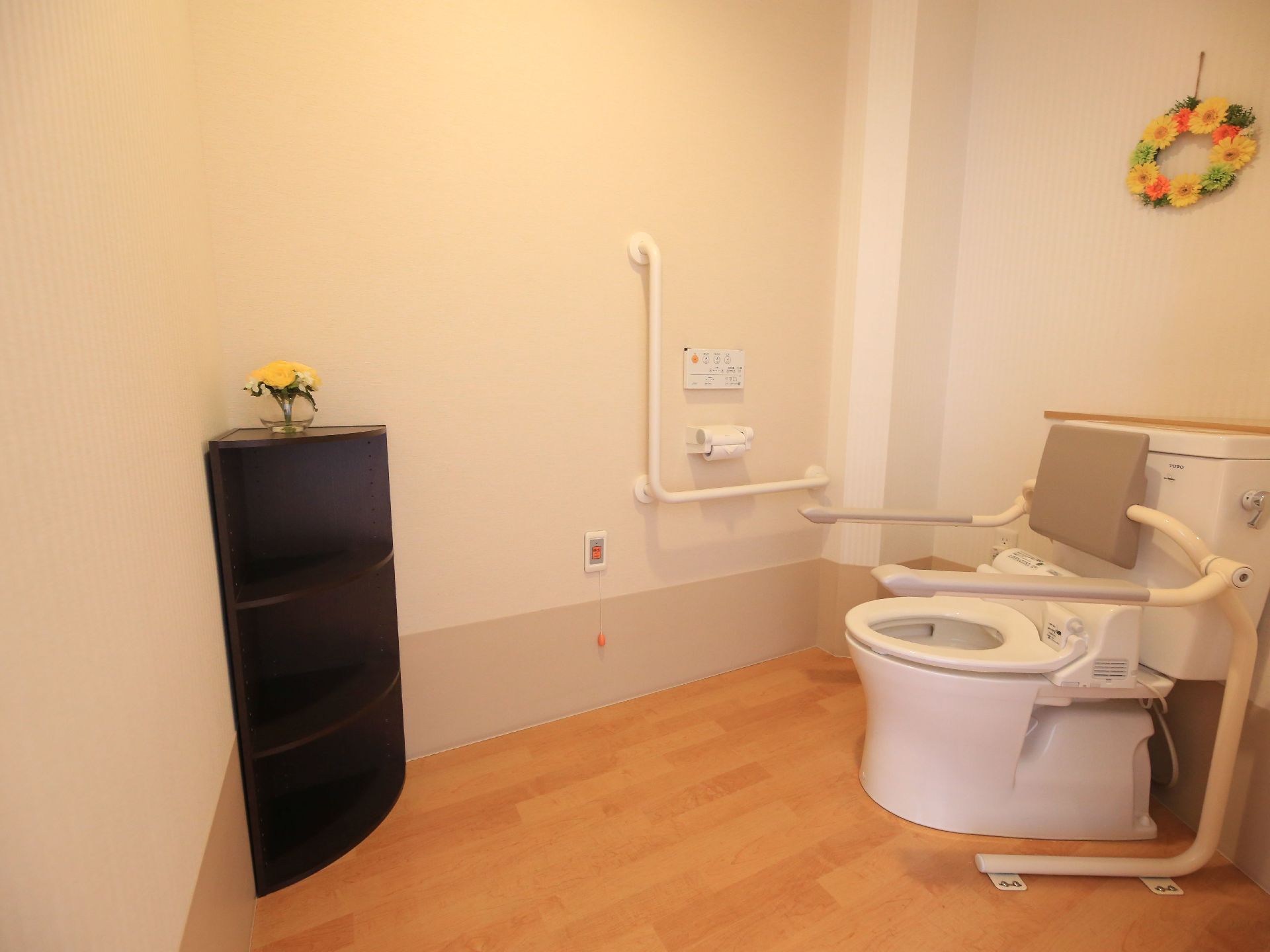 木目調の床を取り入れたトイレスペースである。便座の正面のコーナーに弧を描いた黒の３段ボックスを置き、上に花を飾っている。