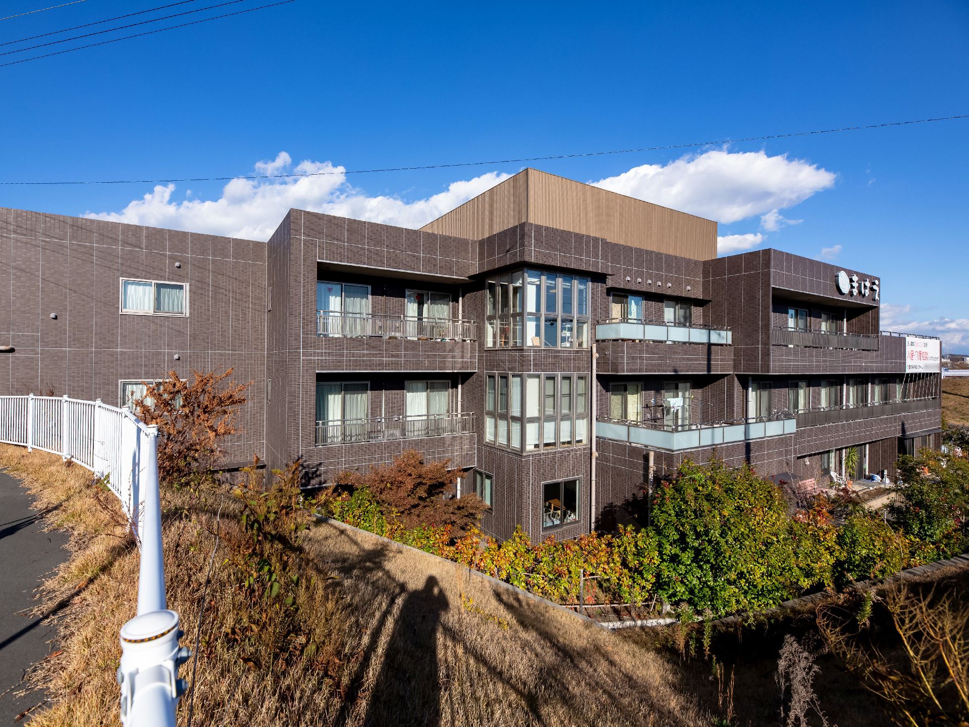ダークブランを基調とした３階建ての外観の施設である。日当り良好な立地で、建物の周りには豊富な植栽を取り入れている。