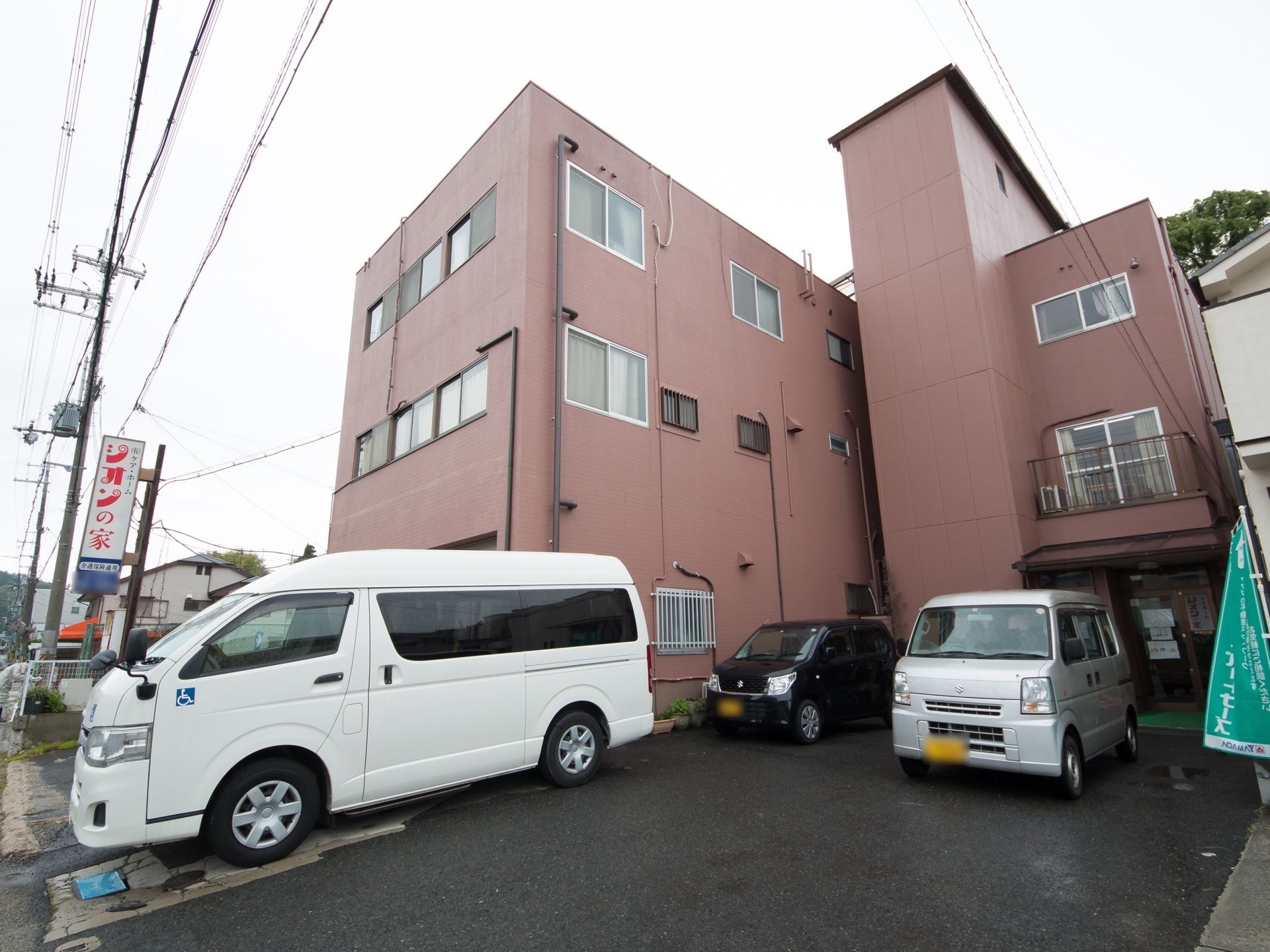 あたたかみのあるバーントオレンジが印象的な、すっきりとした外観の建物である。出入り口の近くには、車が数台停められている。