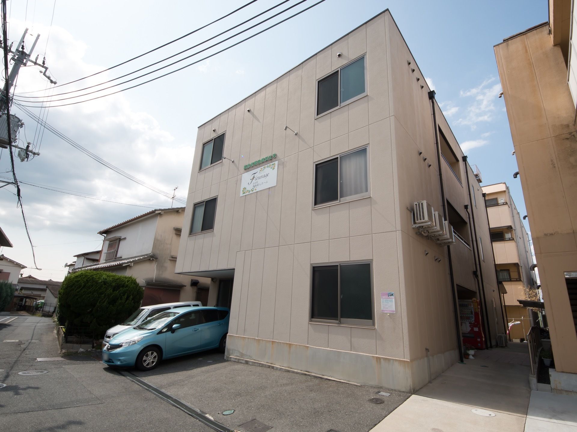 3階建ての建物が見える。1階のベランダ部分には自動販売機が据えつけられている。施設前方に車が2台駐車している。