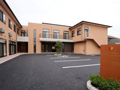広い駐車場のある淡いオレンジ色の外壁の２階建ての施設外観