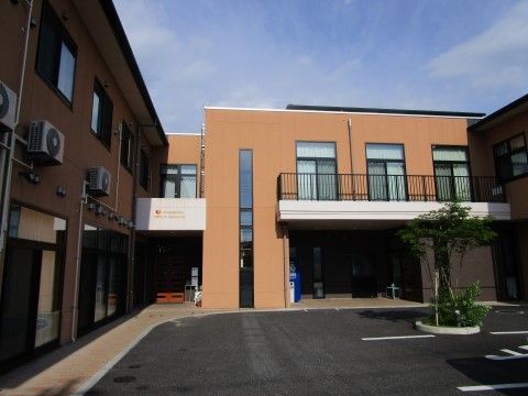 施設は茶色い外観である。1階には青い自動販売機が設置されている。横にはゴミ箱がある。コンクリートの地面の上に植え込みがあり、木が生えている。