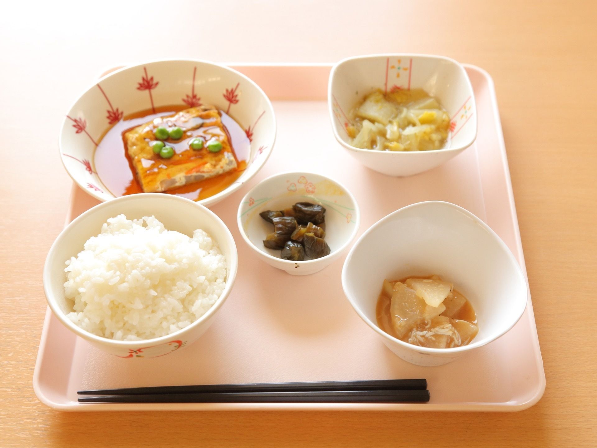食事の提供を行っている。トレイに白いご飯、３種類のおかずと漬物が並んでいる。献立や彩りが工夫されている。