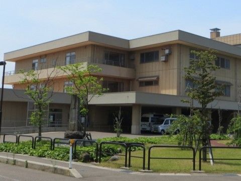 施設の建物の外観である。建物は３階建てで、壁の色はベージュ系である。玄関は屋根付きでスロープがつながっており、駐車場も設けられている。