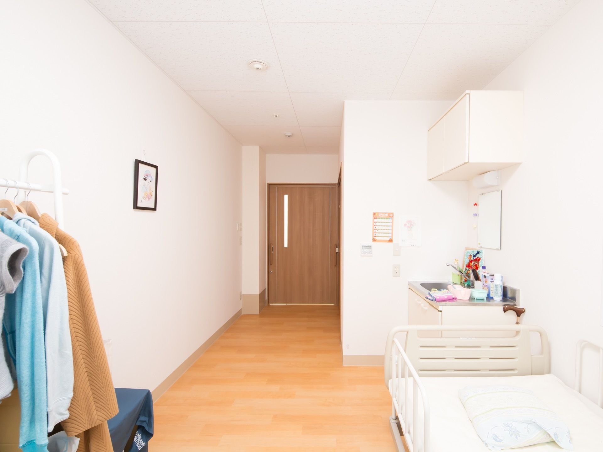 白を基調とした居室である。介護ベッドや流し台、収納が完備されている。壁際に洋服か掛けられたハンガーラックが置かれている。
