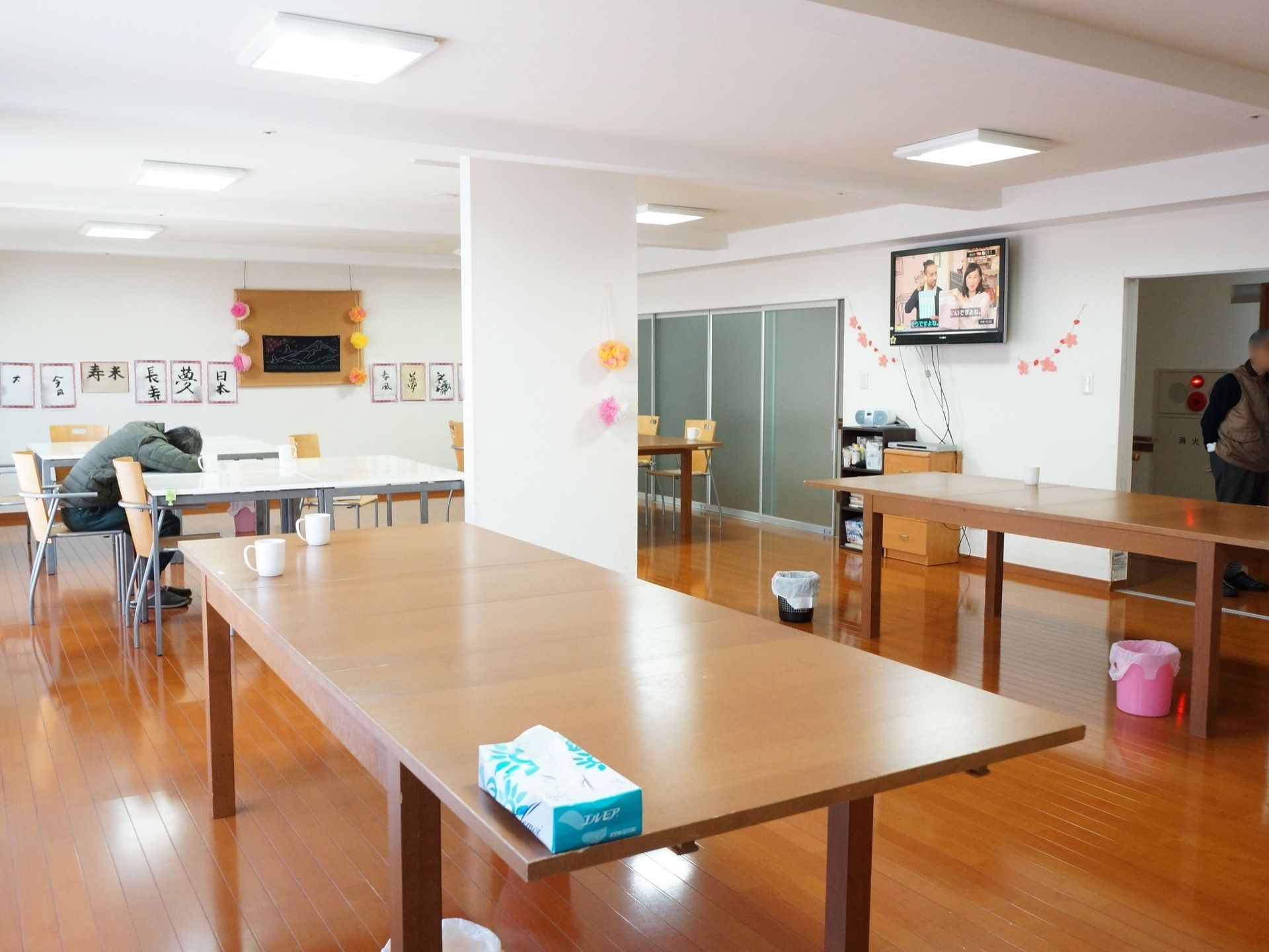 フローリングの部屋に机が数台置いてある。壁には入居者様の習字の作品が飾ってある。テレビを見ながらくつろげる。