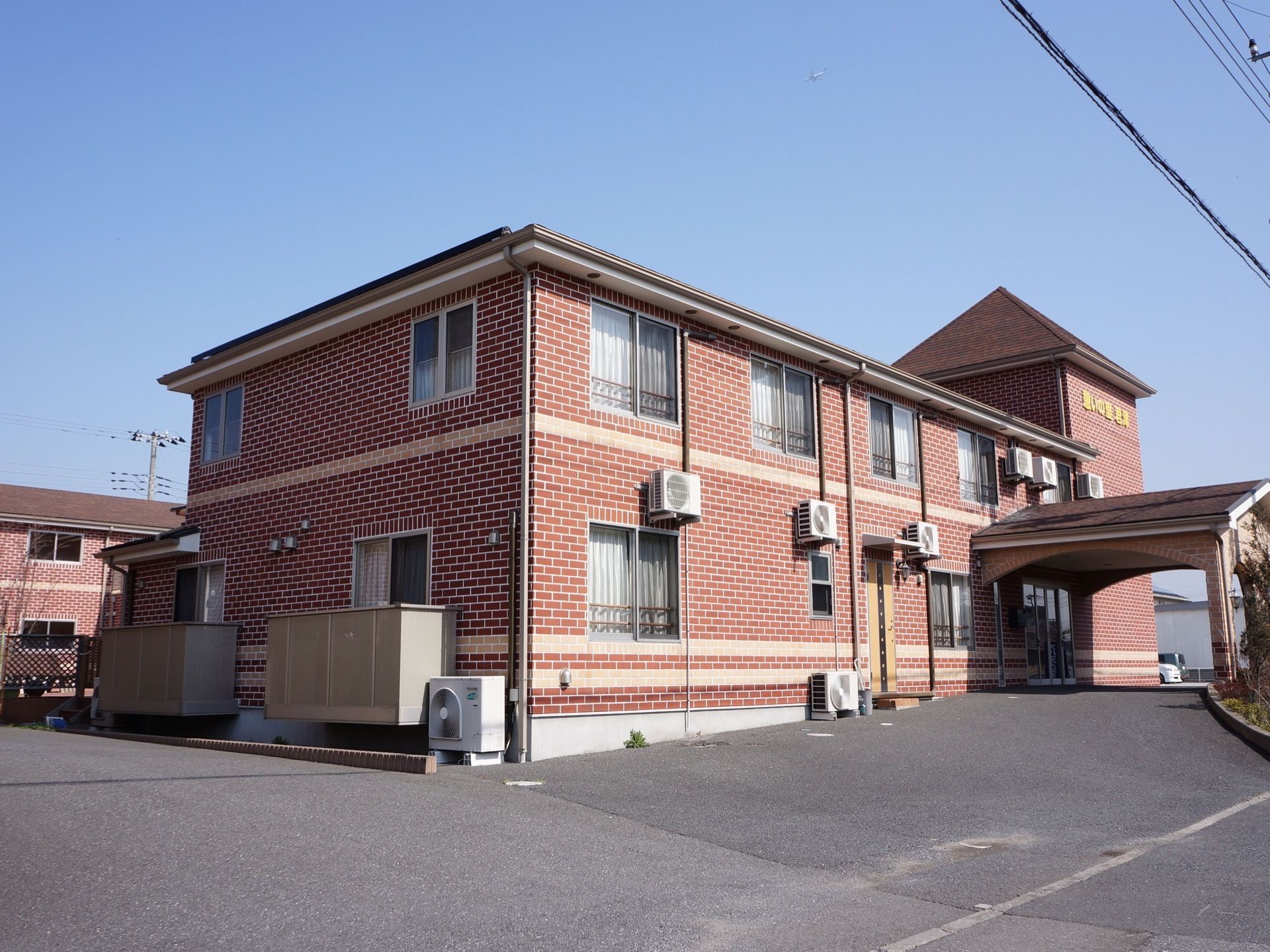 二階建ての建物が見える。外壁はタイル張りになっている。１階の一部のみベランダが付いている。玄関とは別に扉が見える。