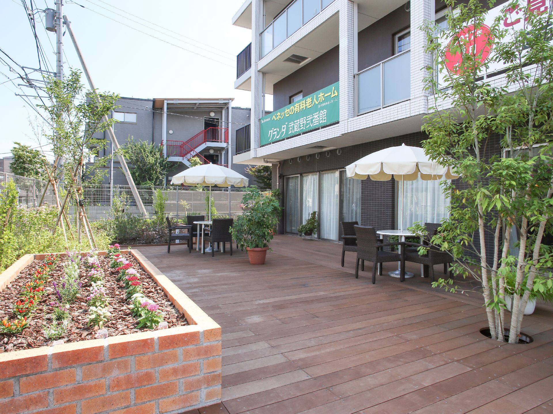 1階にある花壇やグリーンを配したウッドデッキのテラス。パラソル付きのガーデンテーブルとチェア２セットが置いてある。
