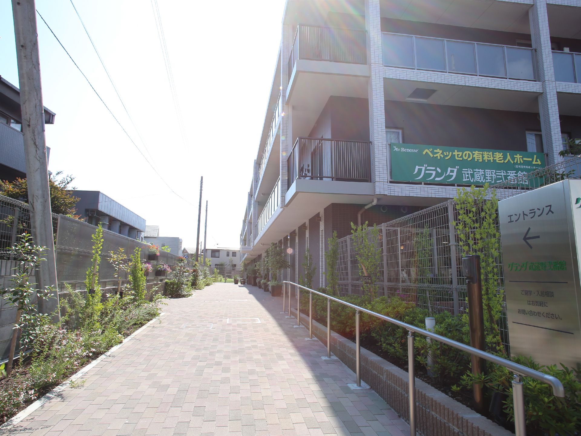 明るいベージュのレンガ敷きの通路。両端には植物とフェンスがある。エントランスの方向を示す看板も設置されている。