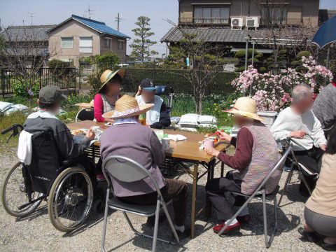 晴れた日に、皆さまが外出先でテーブルを広げ、椅子に腰かけ昼食のお弁当を召し上がっている。スタッフや車椅子をご利用の方の姿も見える。