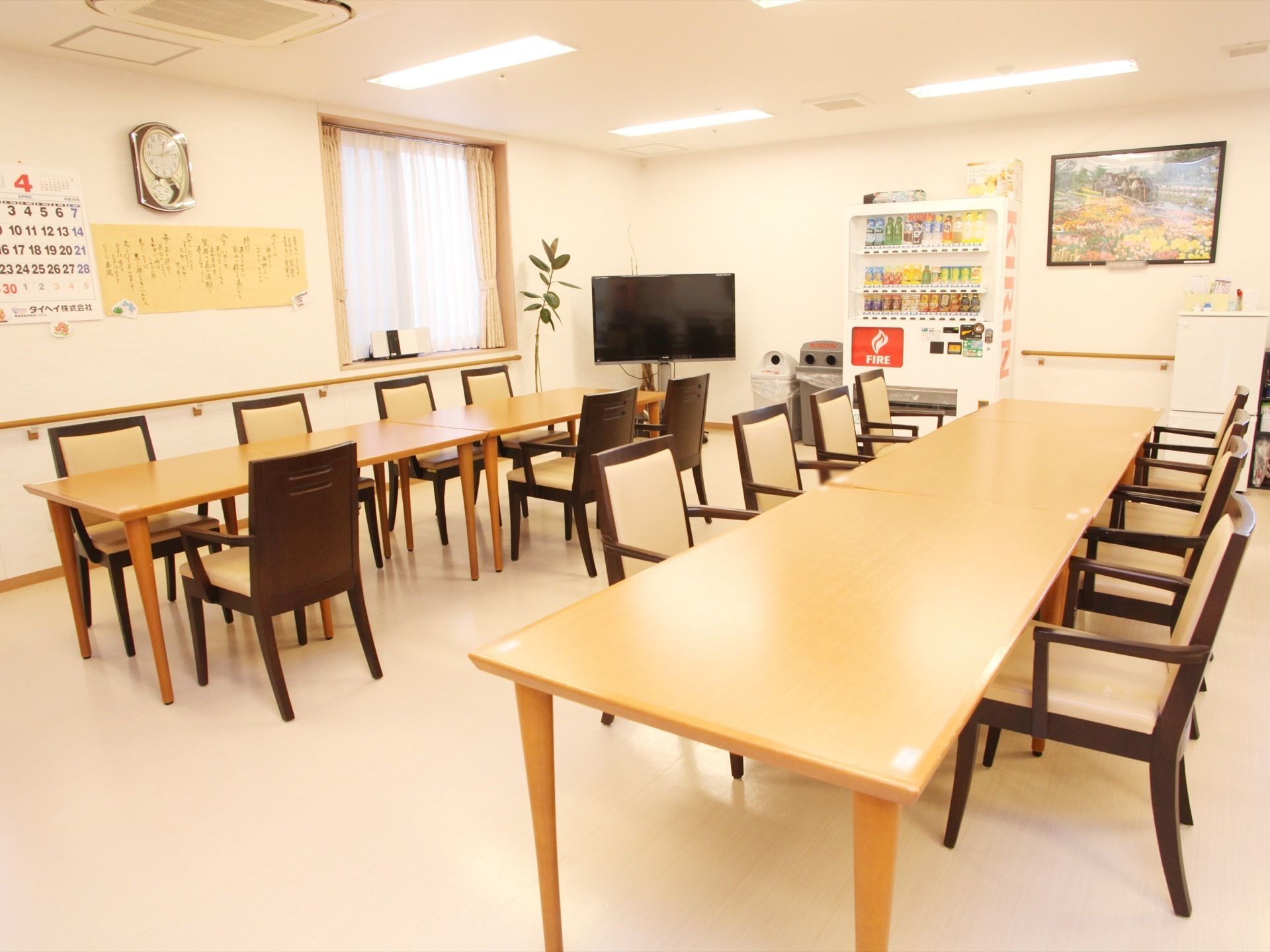 長いテーブルに椅子が複数置かれている。自動販売機やテレビが設置されている。壁には手すりや絵画がある。