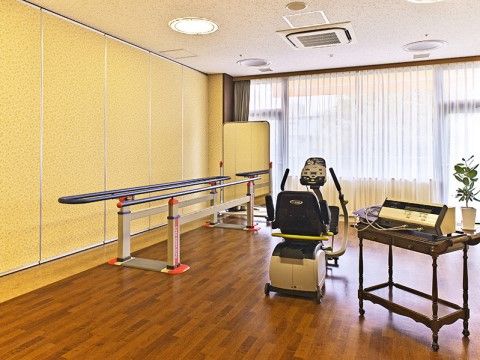 窓がある明るい室内に、平行棒が置いてある。片側が楕円でもう一方が棒になっている。ほかには筋肉トレーニングマシンと、血圧などの測定器がある。