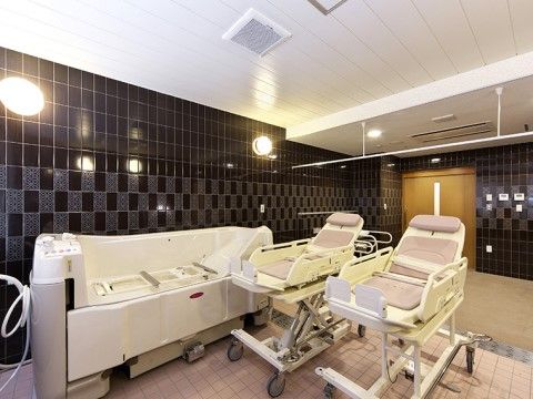 特殊介助浴槽が１台と浴槽に入るためのストレッチャーが２台並んでいる。浴室は壁が黒で床が白色をしている。