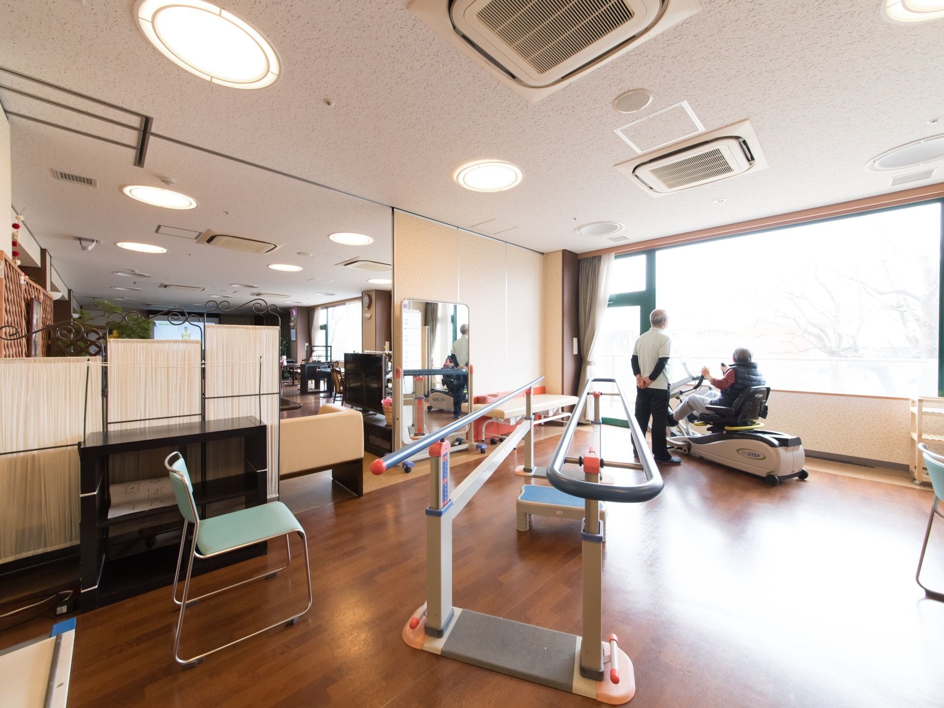 機能訓練中の入居者様とスタッフが見える。大きな窓があり、開放的な空間に感じられる。大きな鏡が設置してある。