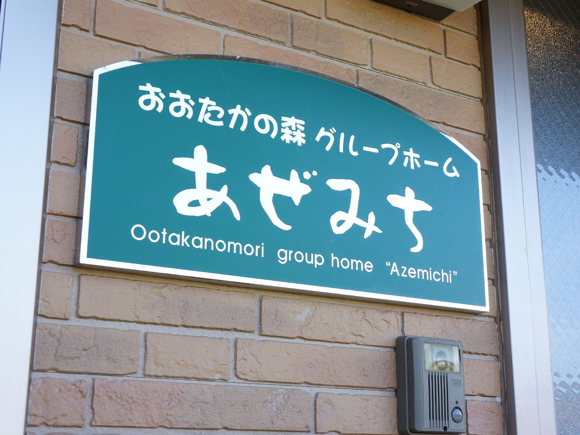 あぜみちと施設名が書かれた看板が設置されている。その下には、呼び出し用のインターフォンもあり使いやすくなっている。