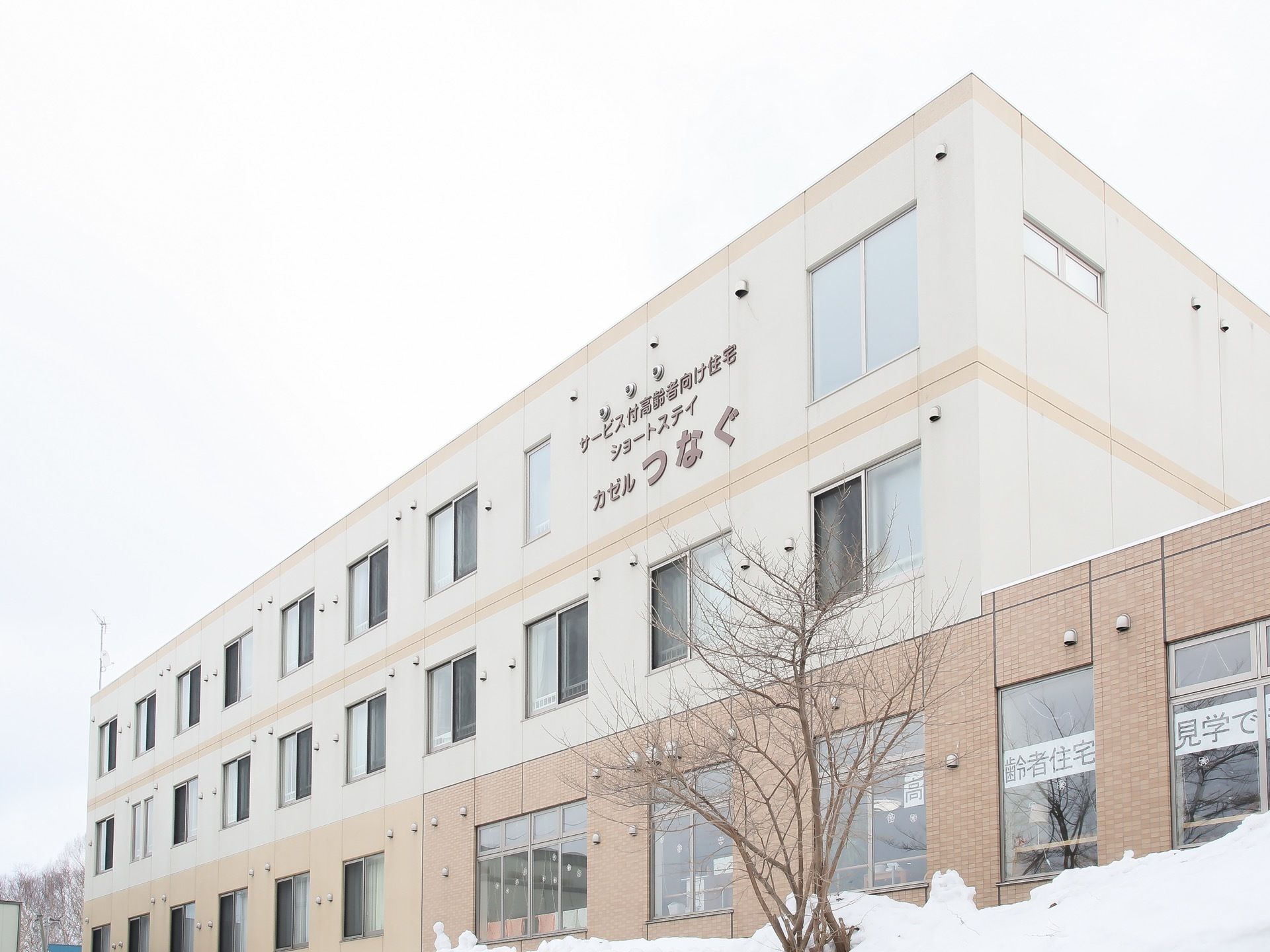 白と茶色を基調とした外壁の建物で、3階の壁面に施設名が書かれている。施設の周囲には樹木などが植えられている。