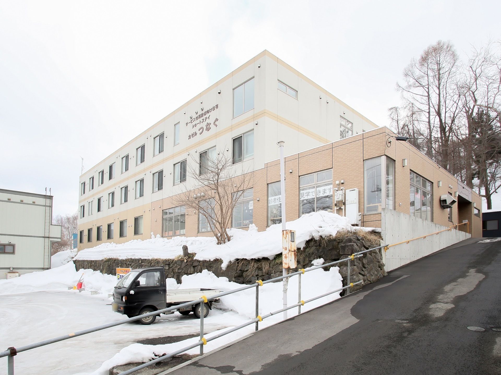 明るい色合いの３階建て施設である。建物の前は、雪が降り積もっている。道路にはガードレールが設けられている。