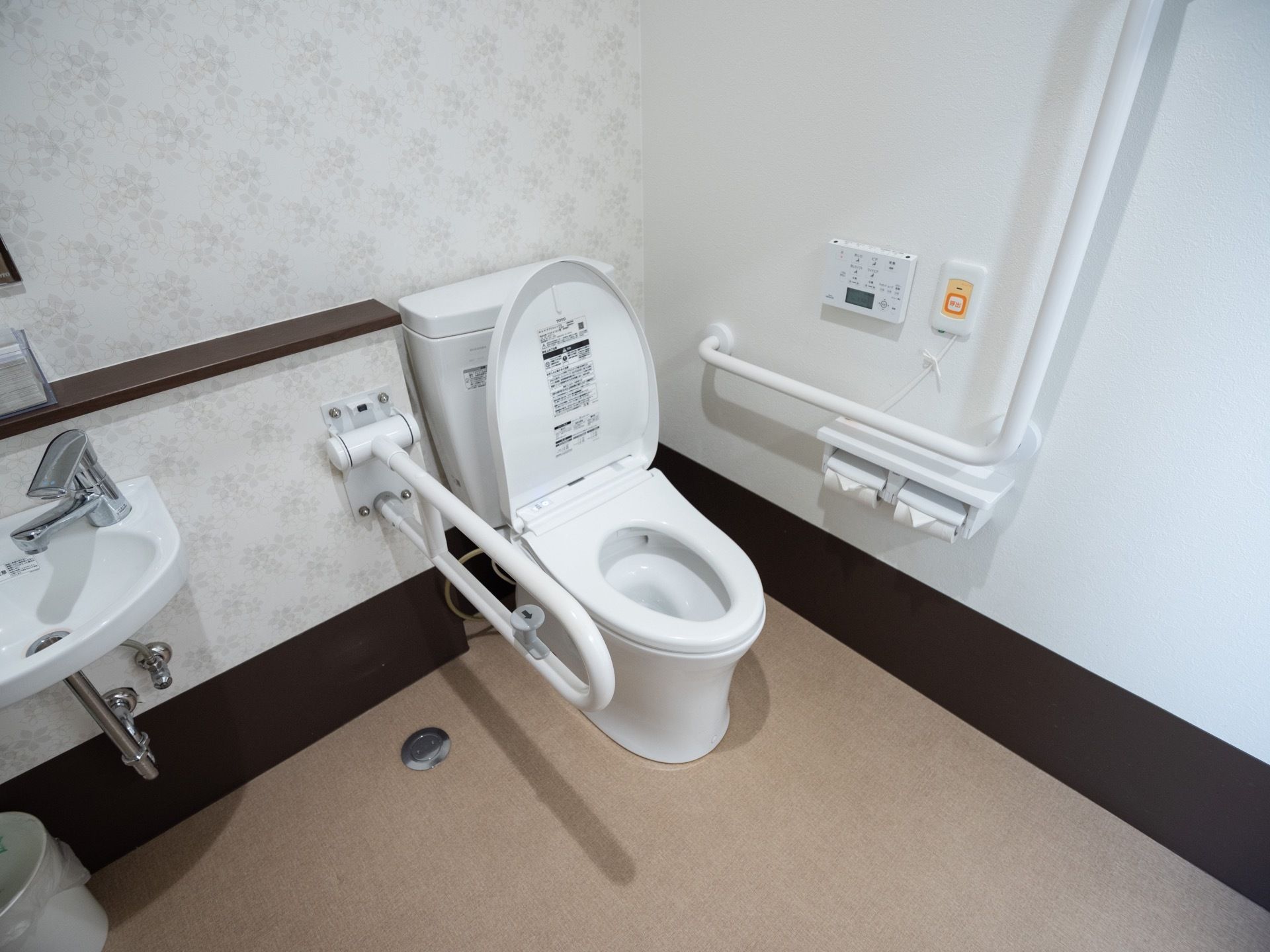白色の洋式便座が備え付けられたトイレである。足元の壁紙は濃い茶色にデザインされている。可動式の手すりもある。