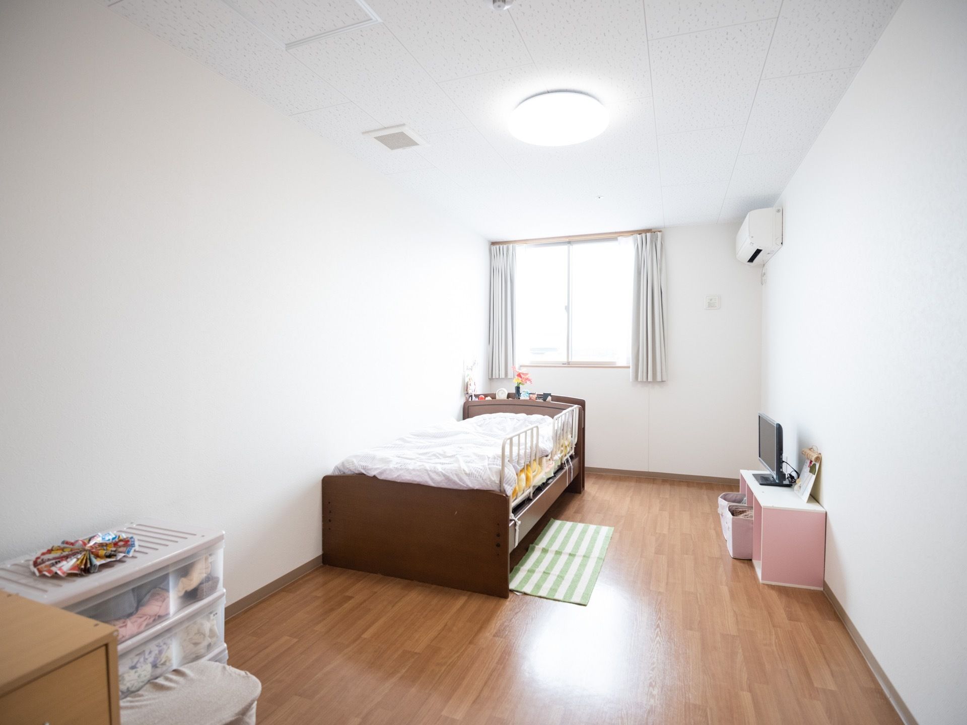 居室は奥行きのある間取りで、シングルベッドや3段ボックス、テレビなどが置かれてる。天井にはシーリングライトが設置されている。