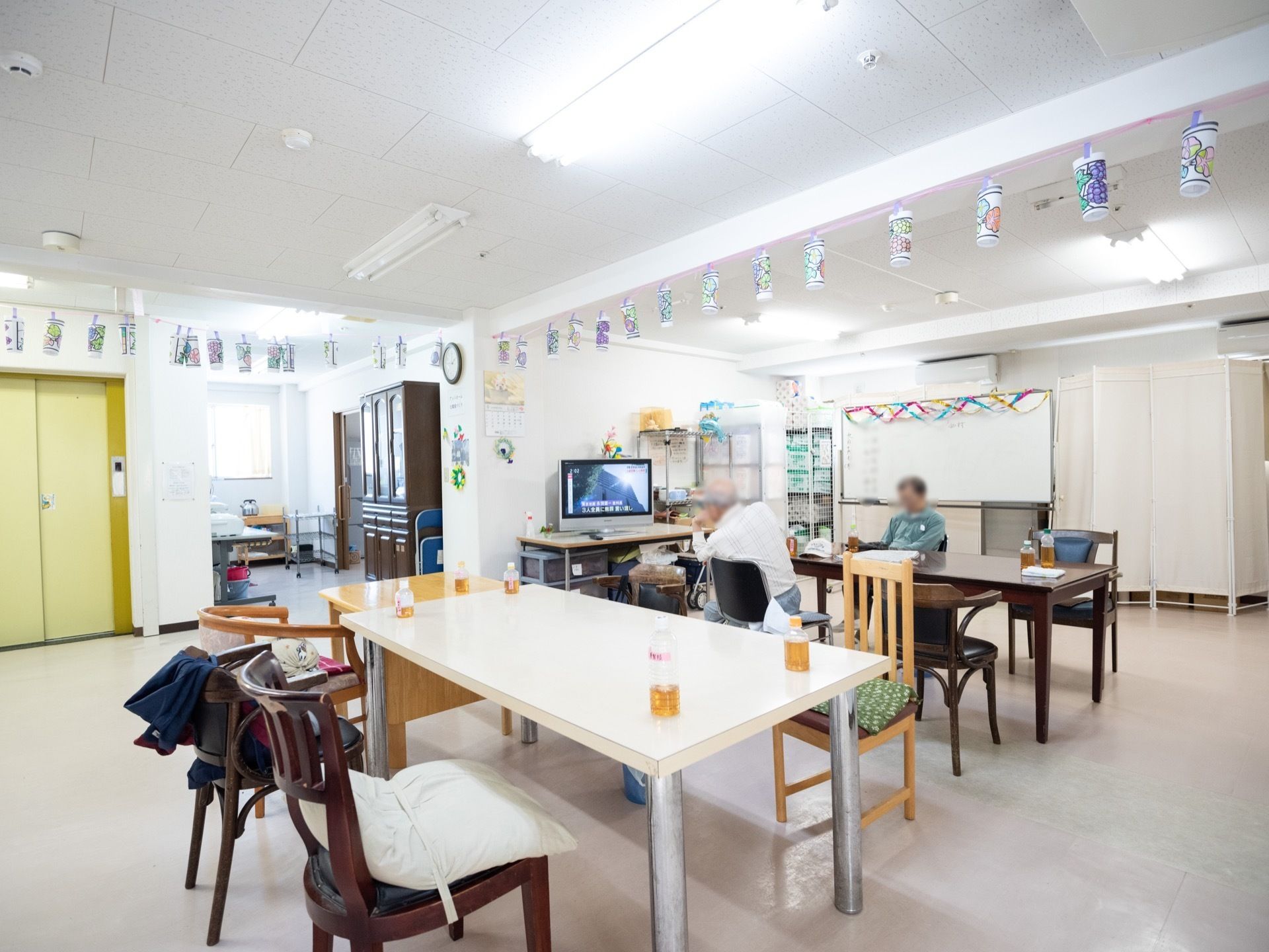 白色を基調とした、共有スペースである。天井に手作りの作品が、たくさん飾られている。テーブルと椅子が置かれている。