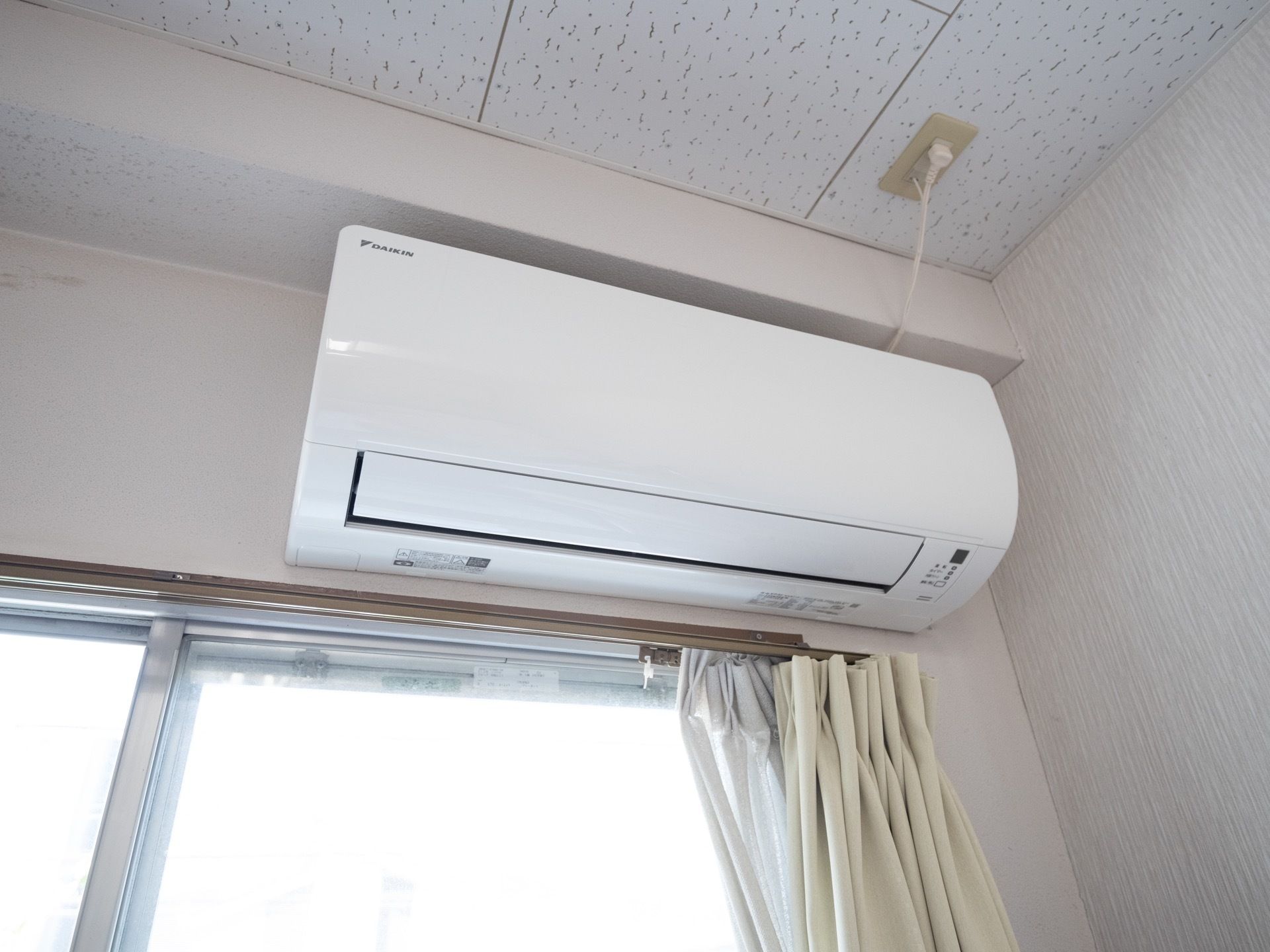 居室に窓が備え付けられ、レースとベージュのカーテンがセットされている。上部には白色のエアコンを設置。