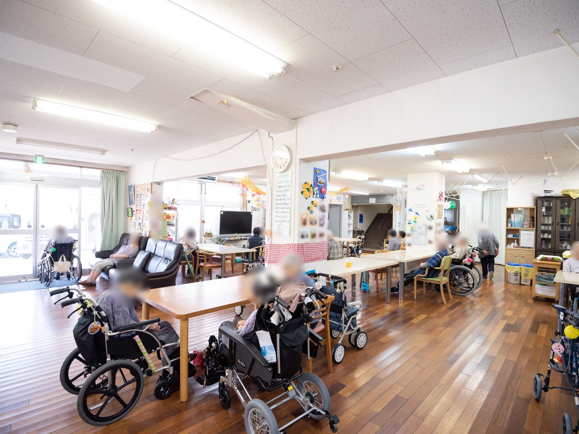 テーブルや椅子が設置された、共有スペース。落ち着いた、茶色のフローリング床である。窓側には、テレビが置かれている。