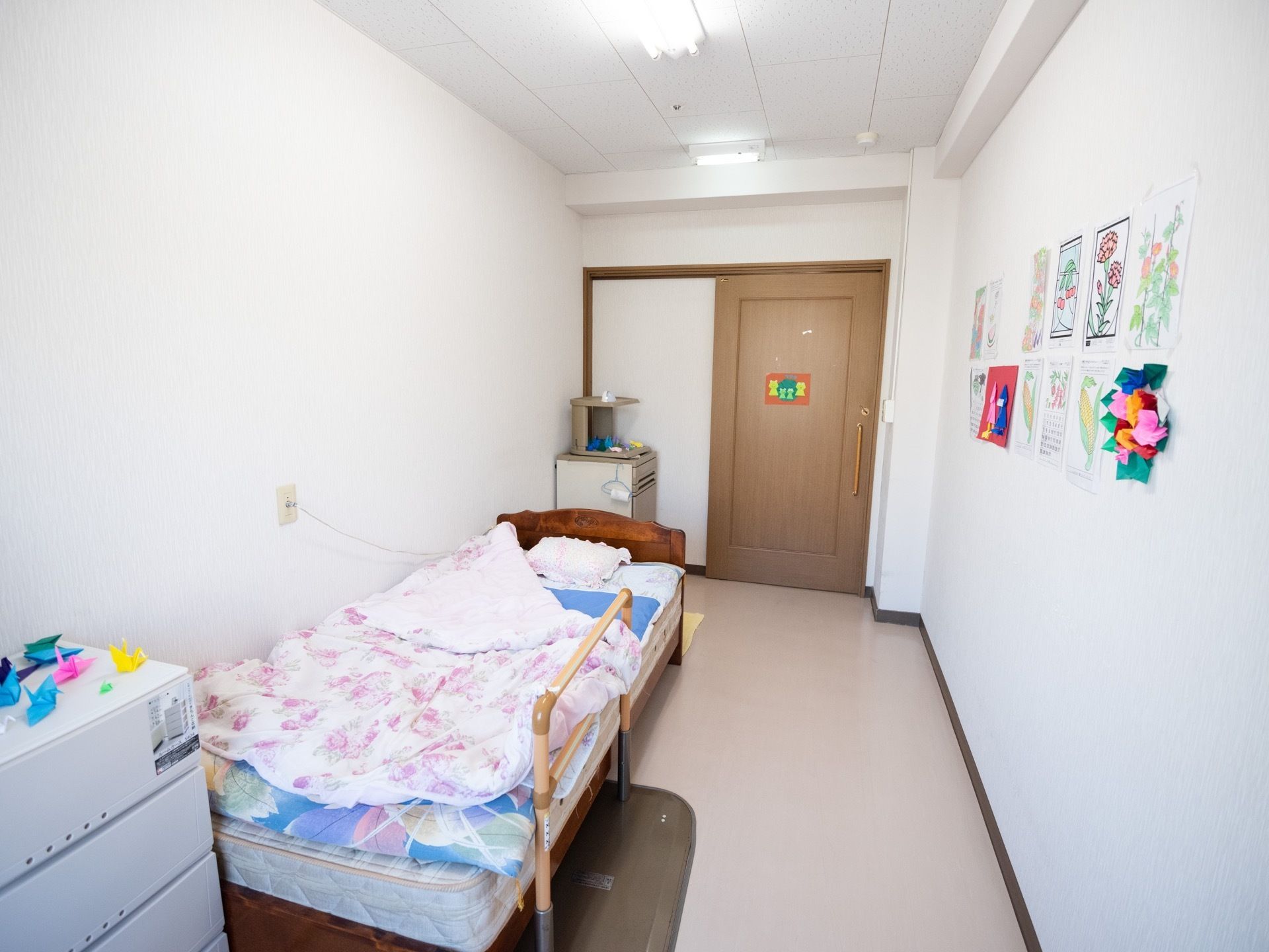 居室の扉は引き戸式である。壁に手作りの作品が飾られ、あたたかい雰囲気。ベッドや収納ケースが置かれている。