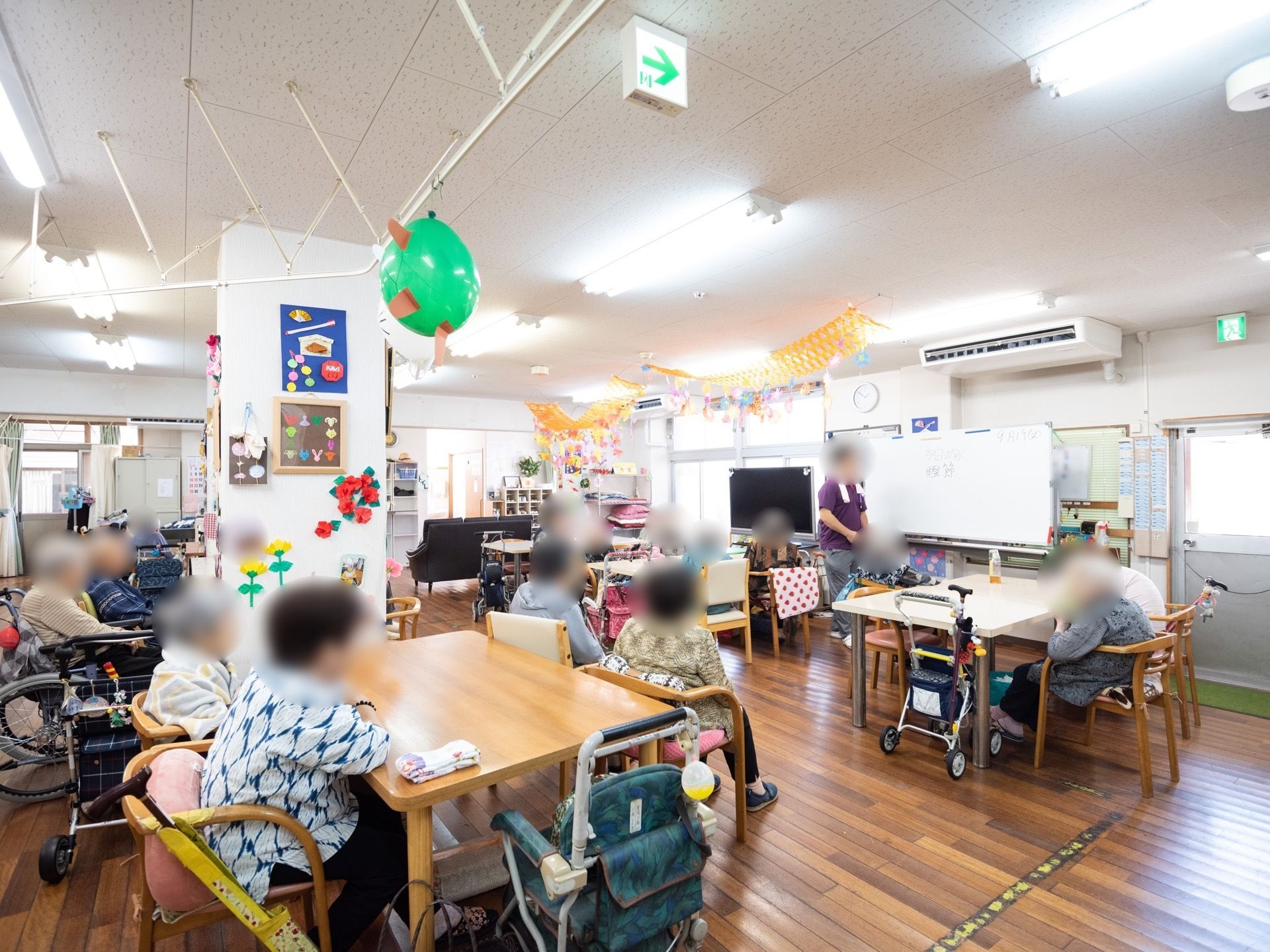 天井に非常口誘導灯が設置された、共有スペースである。スタッフがホワイボード前に立ち、入居者様と交流をおこなっている。